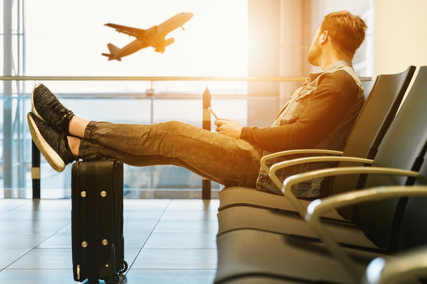 man at airport watching a plane take off with legs on a small travel bag